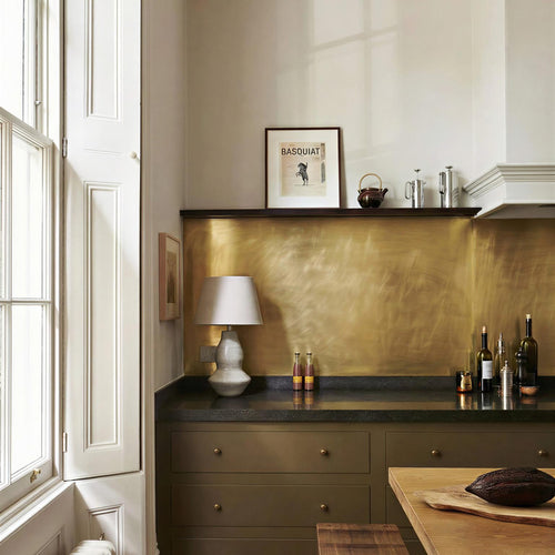 Modern Georgian kitchen with dark stone countertops, olive green cupboards, a floating shelf and brushed brass splashback. There is a classic lamp on a worktop, and a small piece of framed art on the shelf.