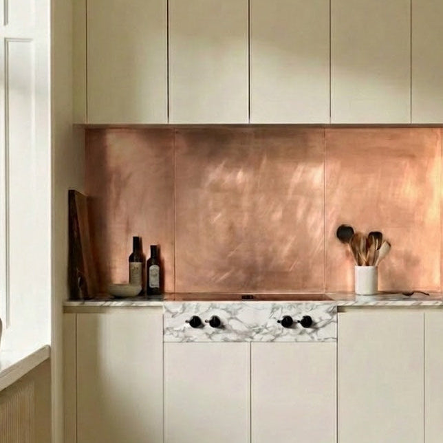 Bespoke brushed rose-gold copper splashback in a modern kitchen with off-white cabinets, a  marble countertop, and herringbone wood floors. 