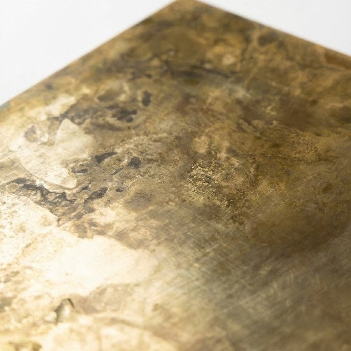 Close-up image of a textured brass surface featuring golden light and clouds of dark patina
