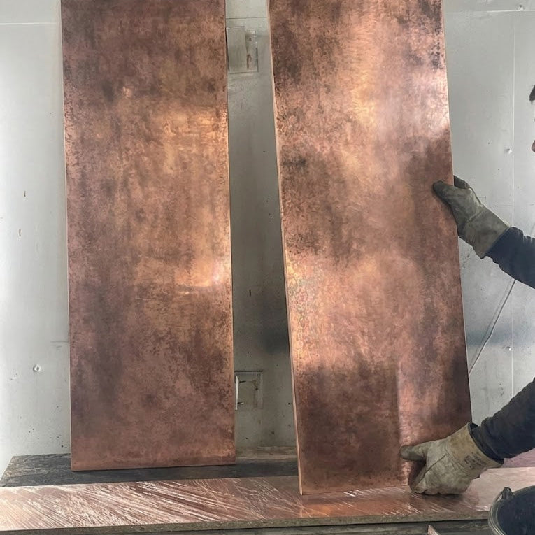 Two large copper panels with dark patina, on a workbench with a person wearing gloves.
