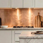 Bespoke rose-gold brushed copper splashback installed behind a gas hob in a luxury white kitchen with marble countertops, waterfall island and minimalist off-white cabinetry. 