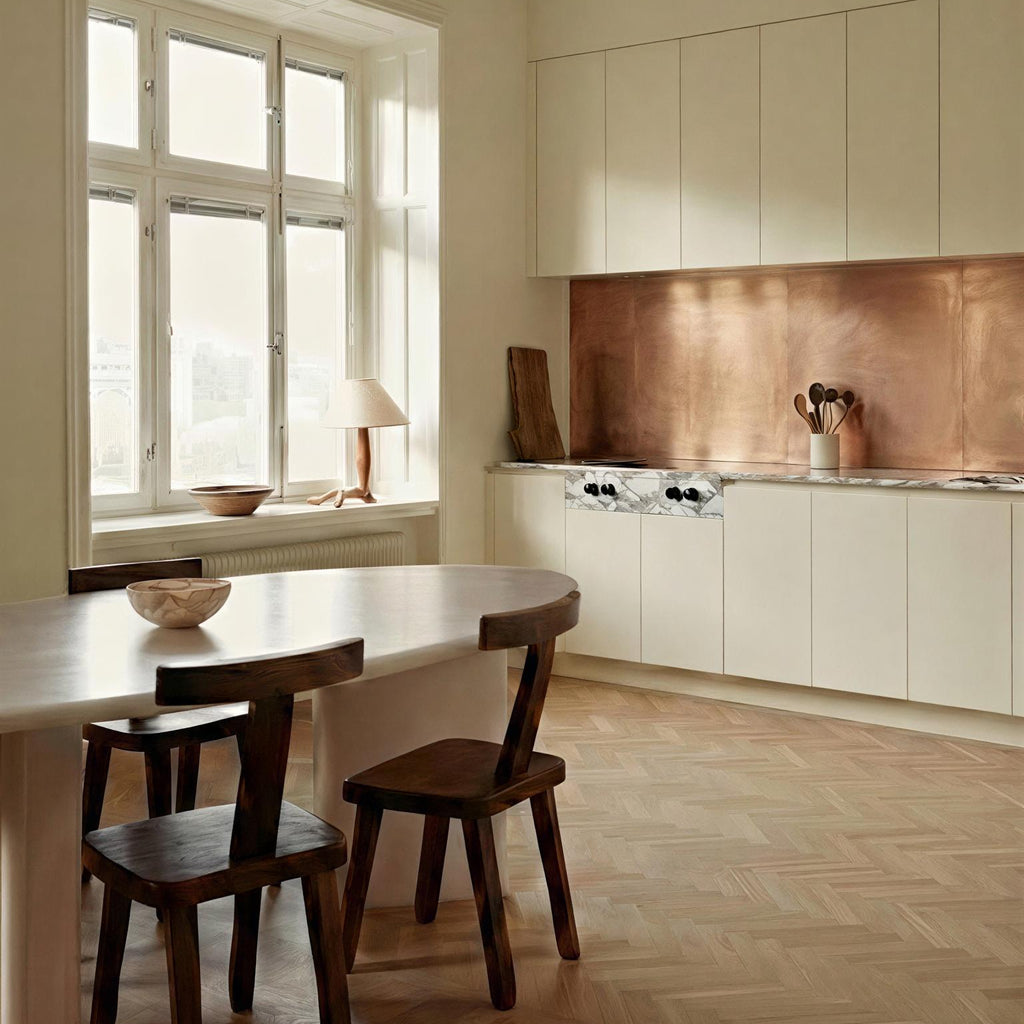 Brushed copper backsplash in a modern kitchen with milk-white flat-panel cabinets. Features include a marble countertop,  herringbone wood floors, a white marble dining table and dark wood chairs. 
