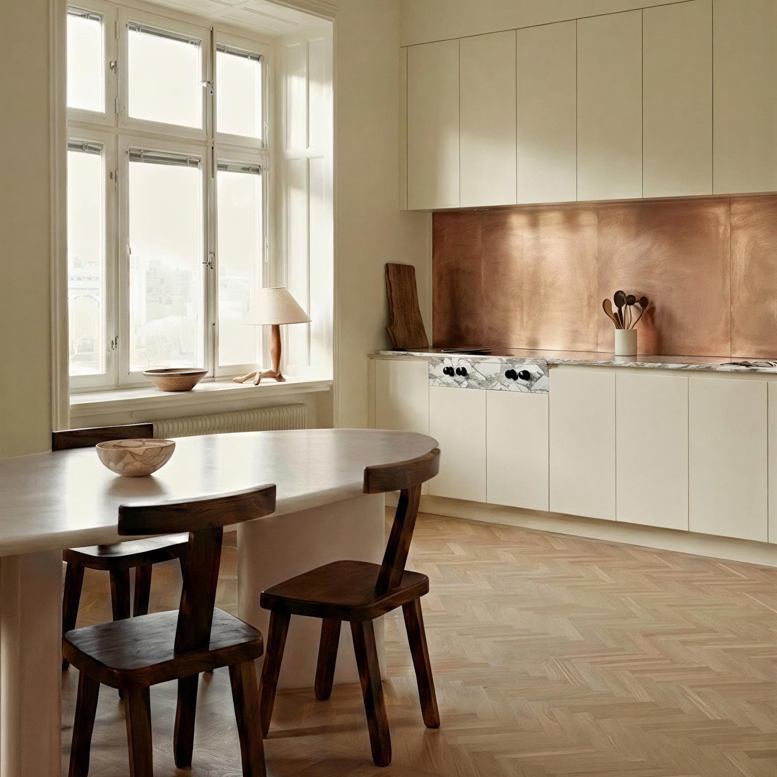 Brushed copper backsplash in a modern kitchen with milk-white flat-panel cabinets. Features include a marble countertop,  herringbone wood floors, a white marble dining table and dark wood chairs. 
