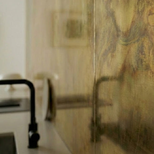 Close-up image of a textured brass splashback with golden light and clouds of dark patina, installed behind a kitchen sink